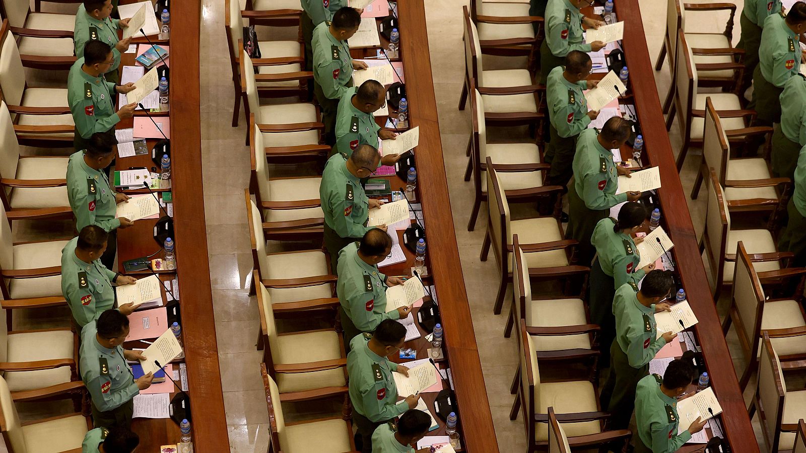 Vista aérea del interior del Parlamento de Naipydó, con representantes uniformados de la Junta Militar de pie, leyendo documentos en sus escaños, cada uno con micrófono y botella de agua.