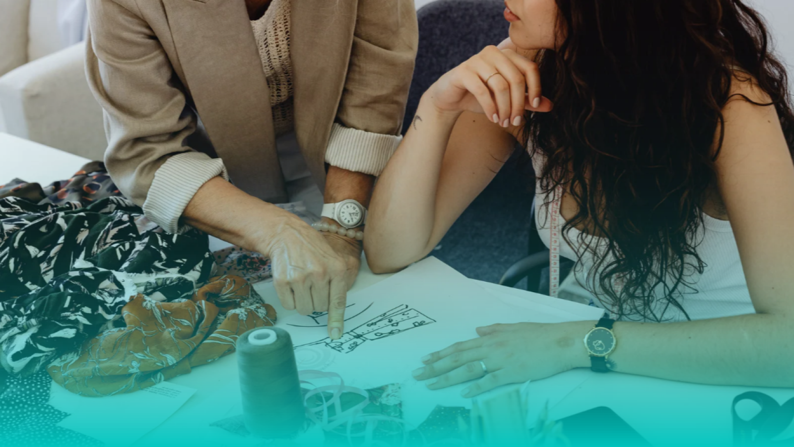 En la imagen, dos mujeres trabajan en un diseño de moda. Una de ellas, con el pelo rizado y camiseta blanca, tiene una cinta métrica. La otra, con chaqueta beige, señala un boceto sobre la mesa, donde hay telas y materiales de costura.