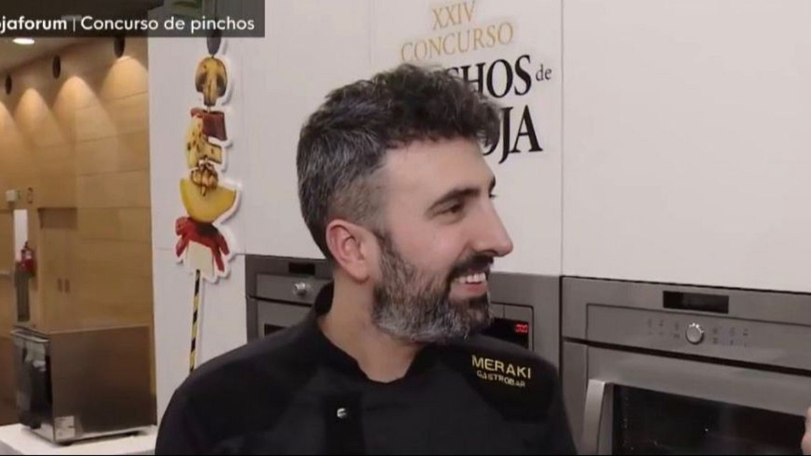 Un hombre con chaqueta de chef negra, sonríe frente a hornos de acero inoxidable. En la imagen, se observa una decoración de pinchos y el nombre de un concurso gastronómico.