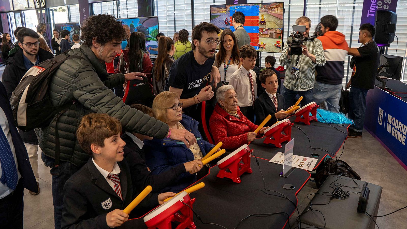 Un grupo de personas de diferentes edades se divierten jugando a un videojuego musical con mandos de tambor en un evento en Madrid, mostrando concentración y disfrute.