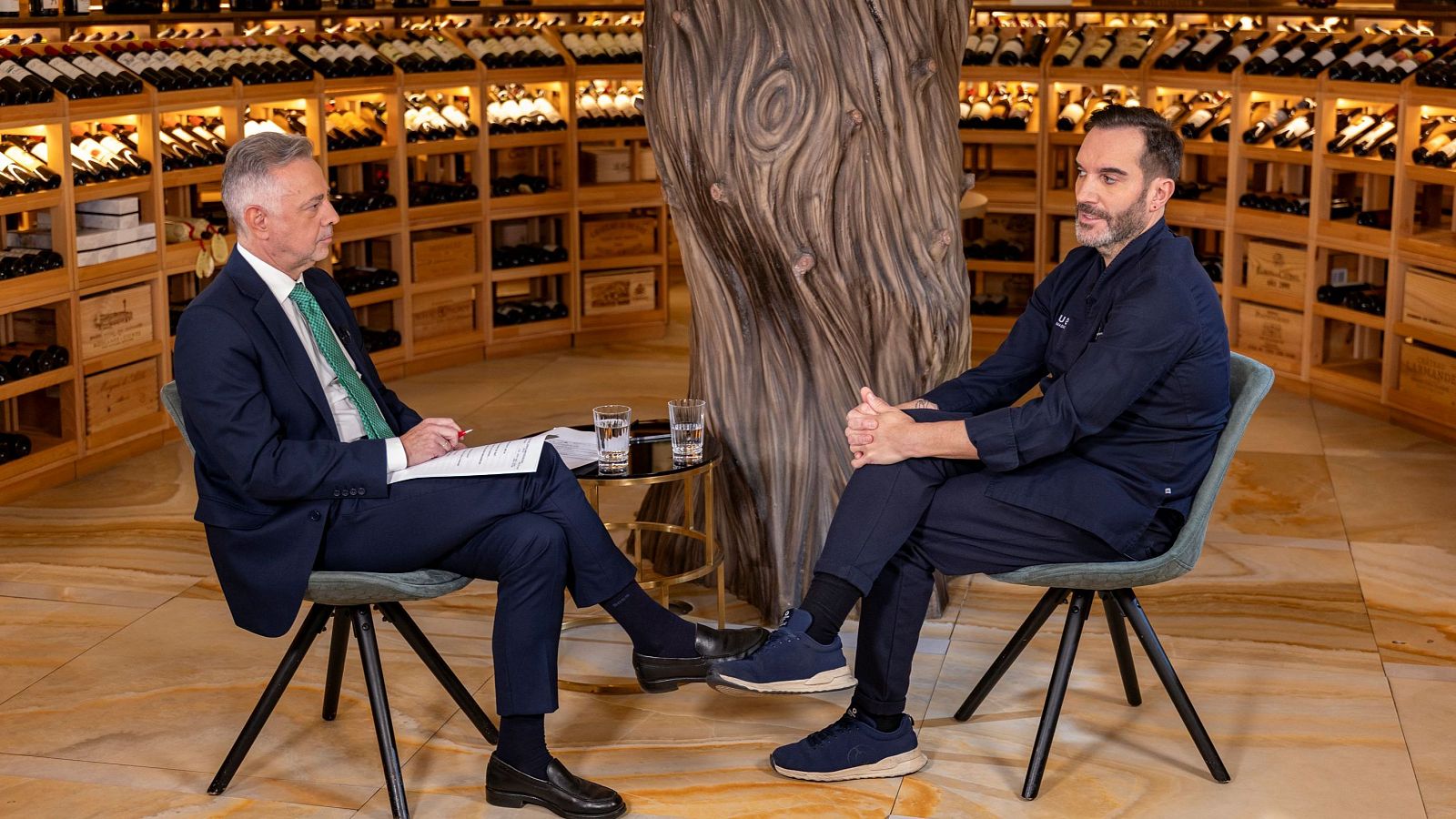 Dos hombres, uno vestido formalmente y otro con uniforme de chef, están sentados frente a frente en una mesa pequeña con vasos de agua. El fondo muestra estanterías de madera llenas de botellas de vino.
