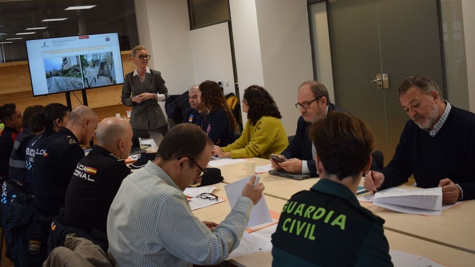 En una sala de reuniones, un grupo de personas uniformadas y una mujer con traje gris participan en una reunión. La mujer parece estar presentando información en una pantalla, mientras los asistentes toman notas.