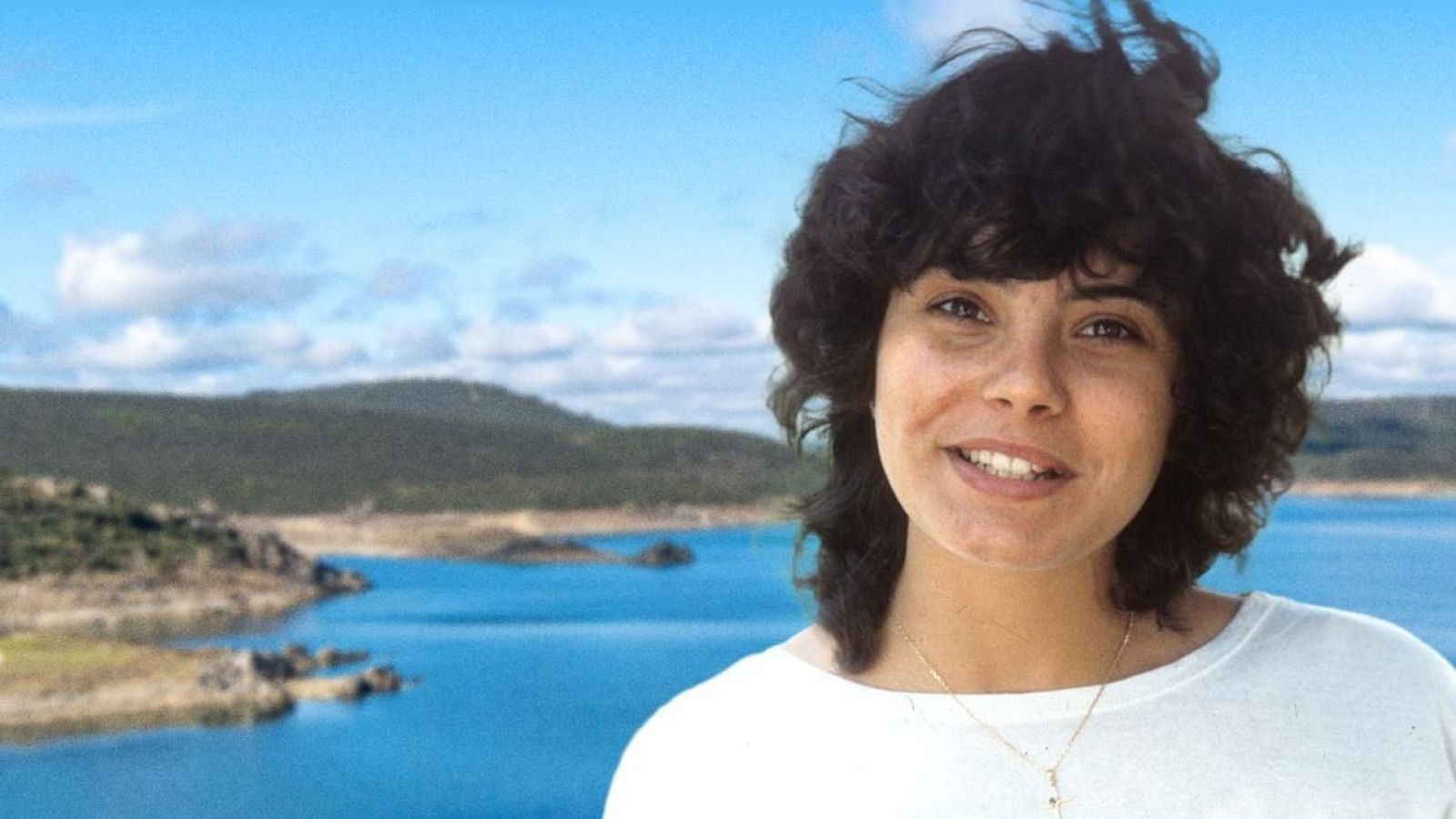 Una mujer de piel clara, con cabello corto y rizado, sonríe a la cámara. Lleva una camiseta blanca y un collar dorado, con un paisaje natural de fondo.