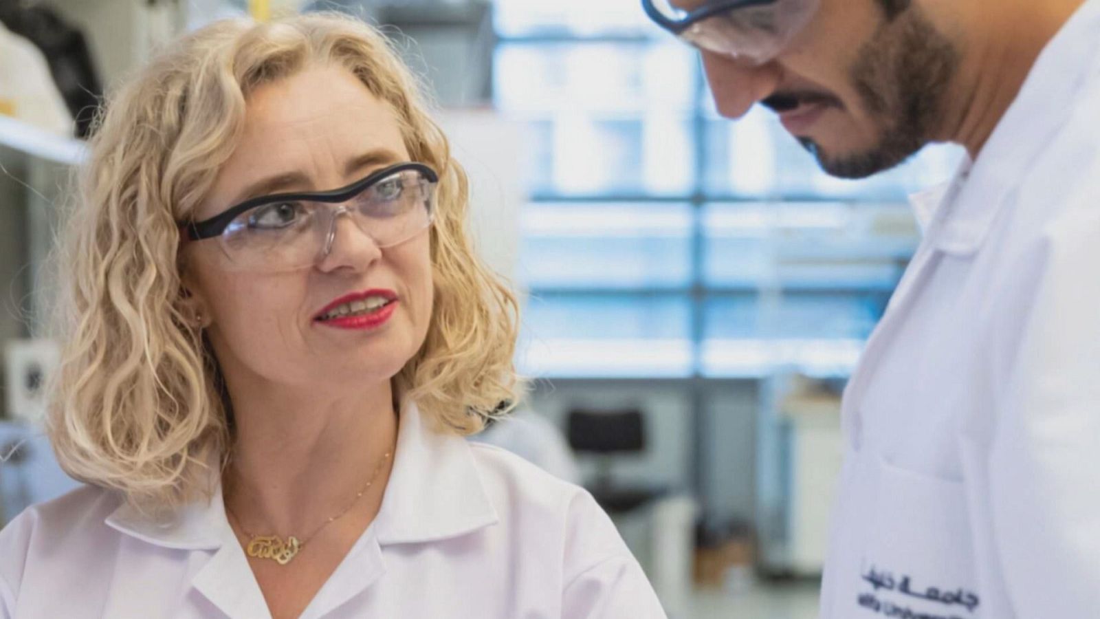 Dos científicos, una mujer con labial rojo y un hombre barbudo, posan en un laboratorio. Ambos visten batas blancas. El hombre tiene una bata con escritura en árabe.