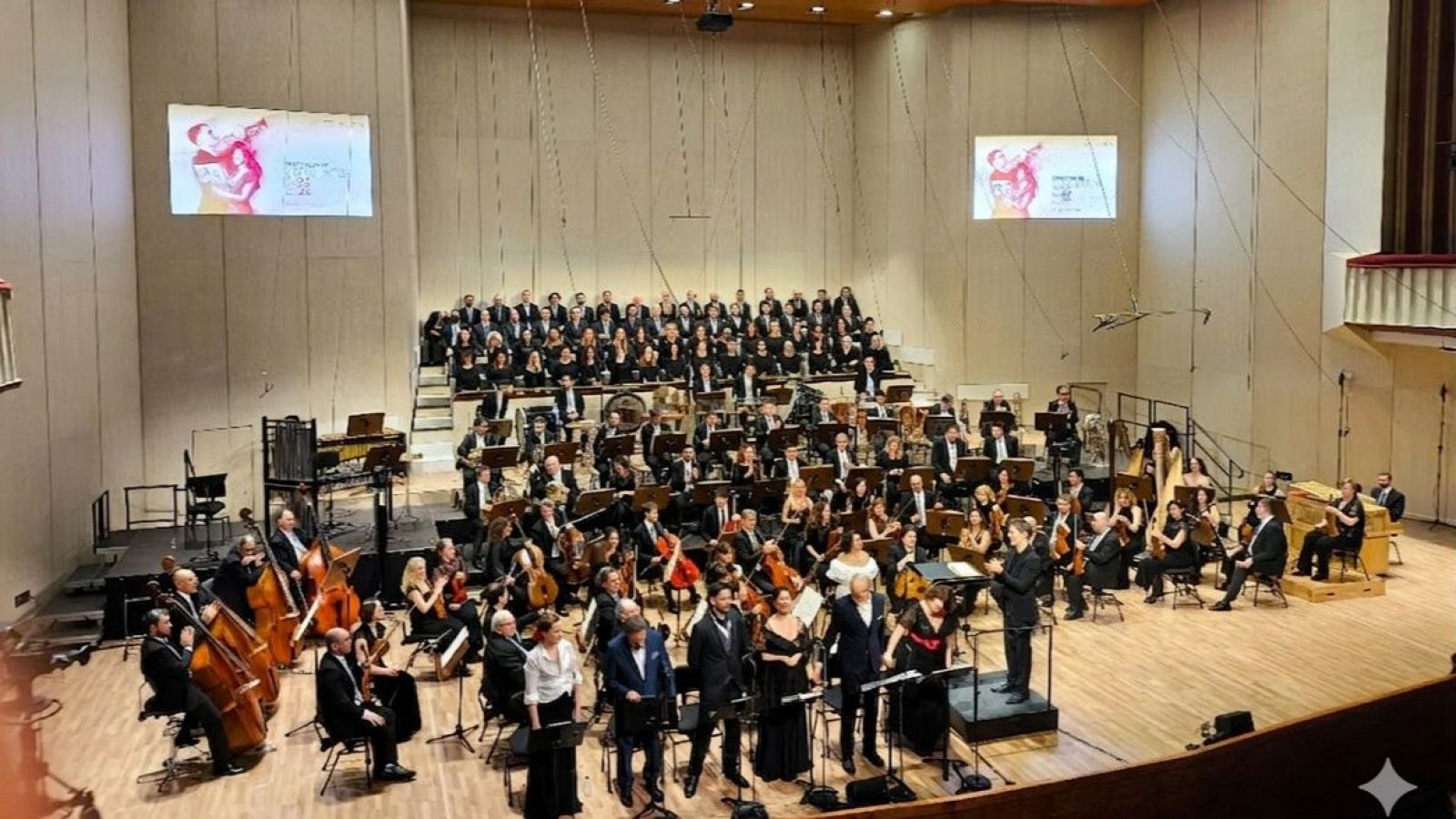Una orquesta y un coro, ambos vestidos formalmente, actúan en un escenario iluminado. Los músicos de la orquesta tocan instrumentos de cuerda, mientras que el director dirige desde un podio. El coro se encuentra en una plataforma elevada.