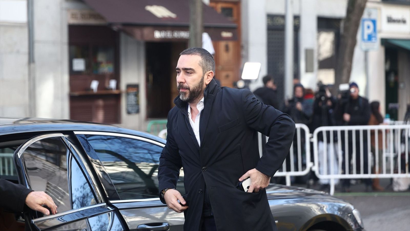 Víctor de Aldama, con abrigo oscuro y camisa blanca, desciende de un coche frente a un edificio, posiblemente un tribunal. Se aprecia la presencia de cámaras y otras personas en el entorno.