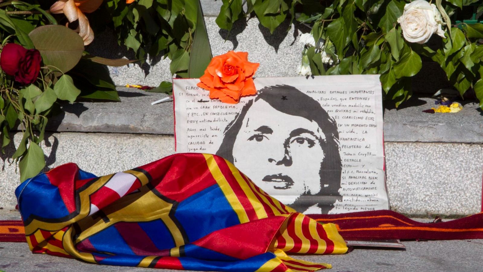 Un memorial en el Camp Nou exhibe una bandera del club y un retrato en blanco y negro, junto a un texto manuscrito y flores de varios colores.