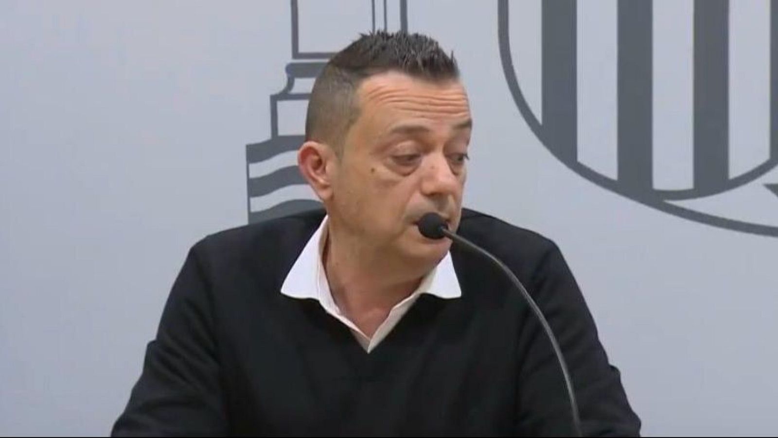Un hombre con cabello oscuro y corte moderno, con expresión seria, habla por un micrófono. Viste camisa blanca y jersey negro, con un fondo gris y un logotipo.