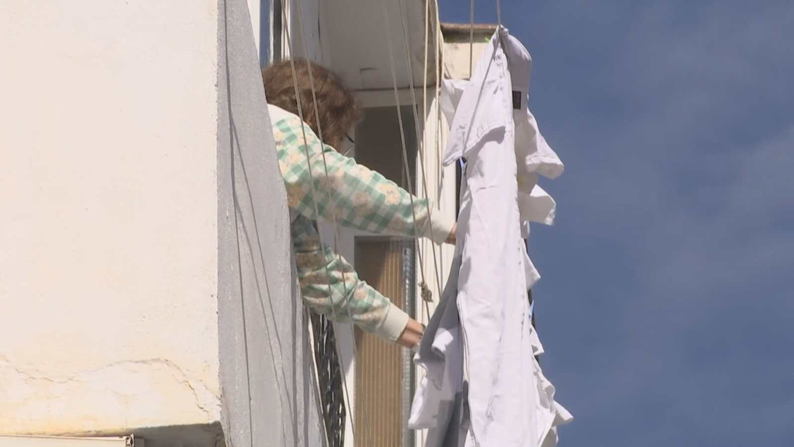 Una mujer con camisa de cuadros extiende ropa en un tendedero exterior, fijado a una pared blanca, con el cielo azul visible.