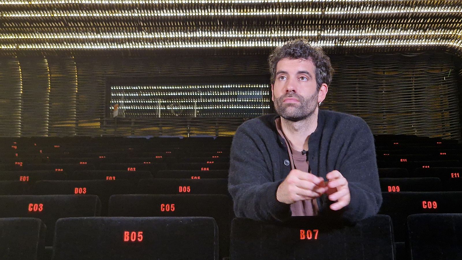 En una sala de cine, un hombre con barba y pelo rizado, vestido con chaqueta oscura, está sentado en una butaca con un número rojo. Se observan más butacas negras con números rojos en la sala.