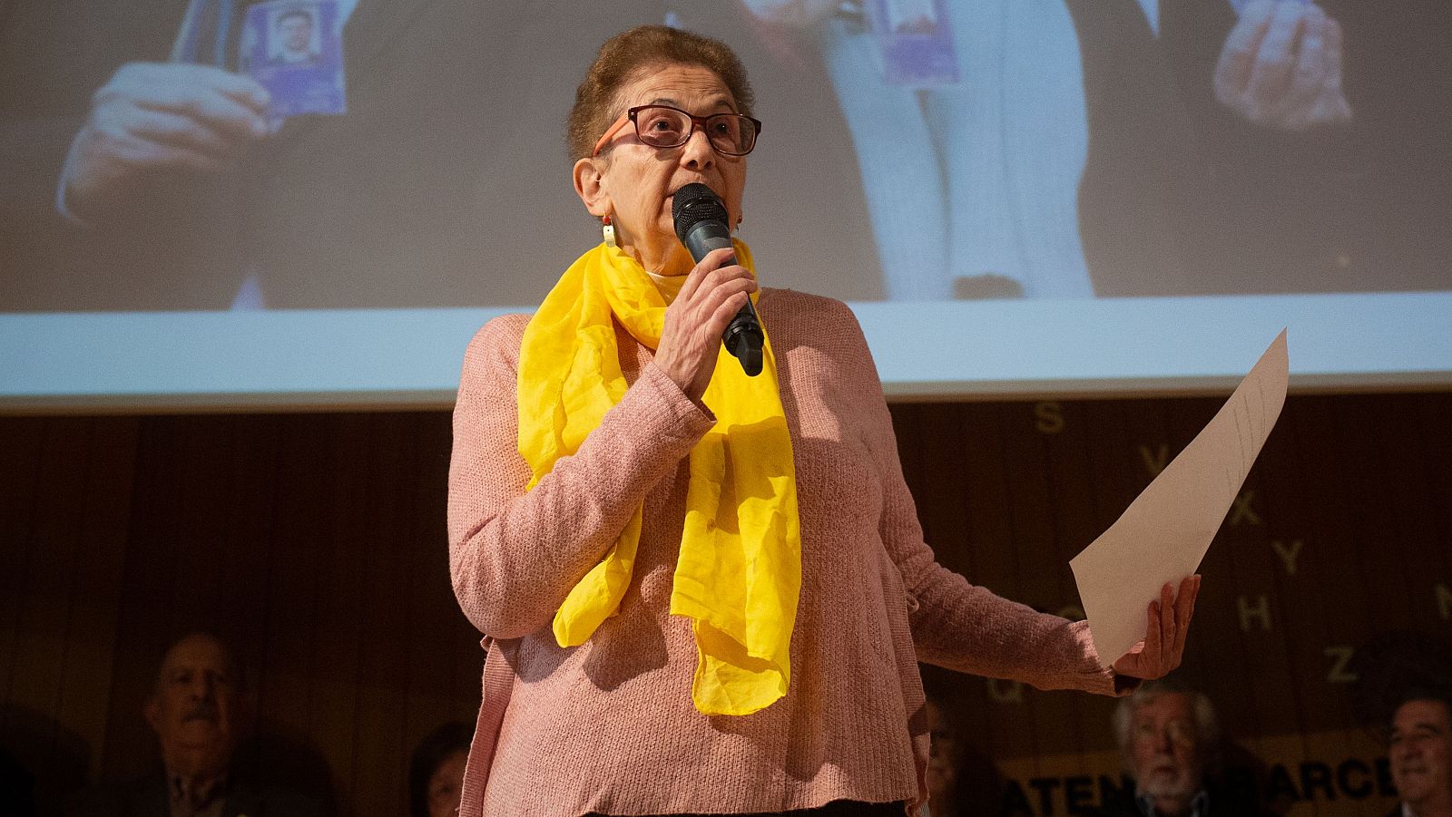 Una mujer mayor, con gafas y pelo rizado, sostiene un micrófono y un papel, posiblemente hablando en público. Viste una blusa rosa y una bufanda amarilla, y se encuentra frente a una audiencia.