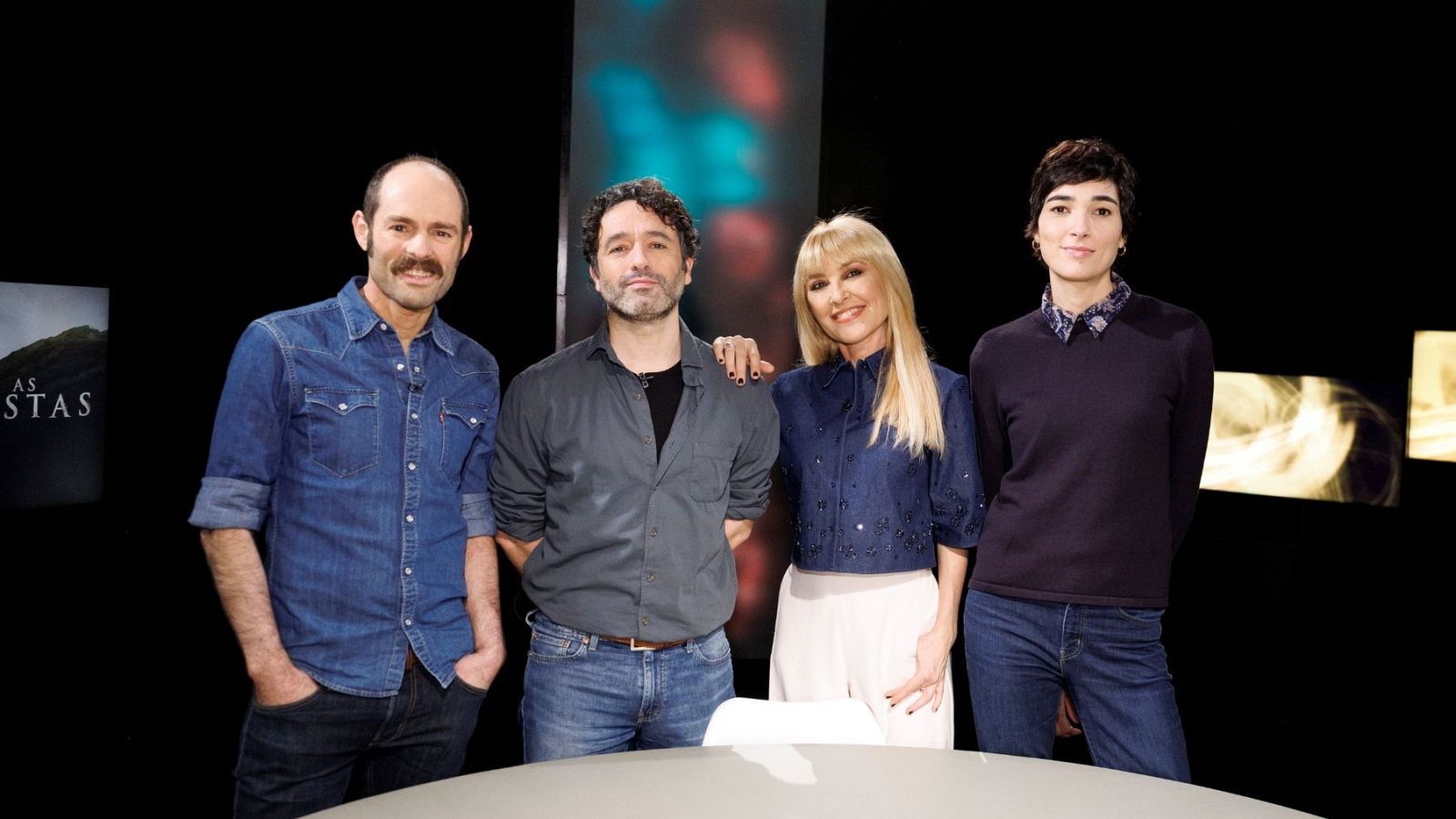 Cuatro personas posan en un estudio con fondo negro: un hombre con camisa vaquera, otro con camisa gris, una mujer con blusa vaquera y falda blanca, y otra con jersey azul oscuro. Al fondo, elementos decorativos abstractos.