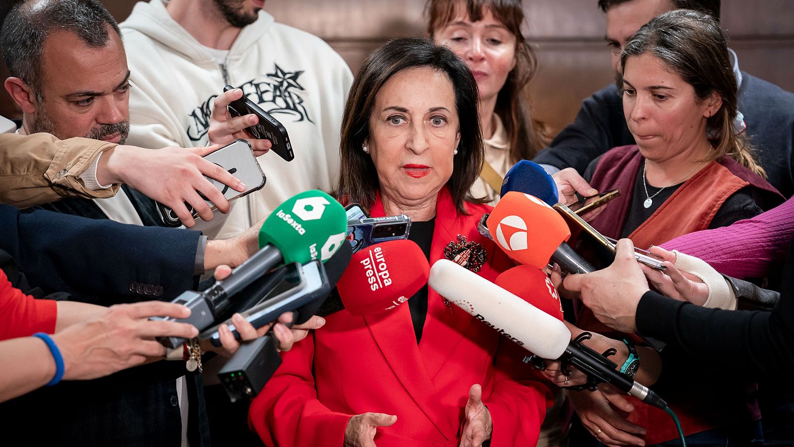 La Ministra de Defensa, Margarita Robles, atiende a los medios de comunicación, rodeada de micrófonos con logotipos de varios medios, con una chaqueta roja y una expresión seria.