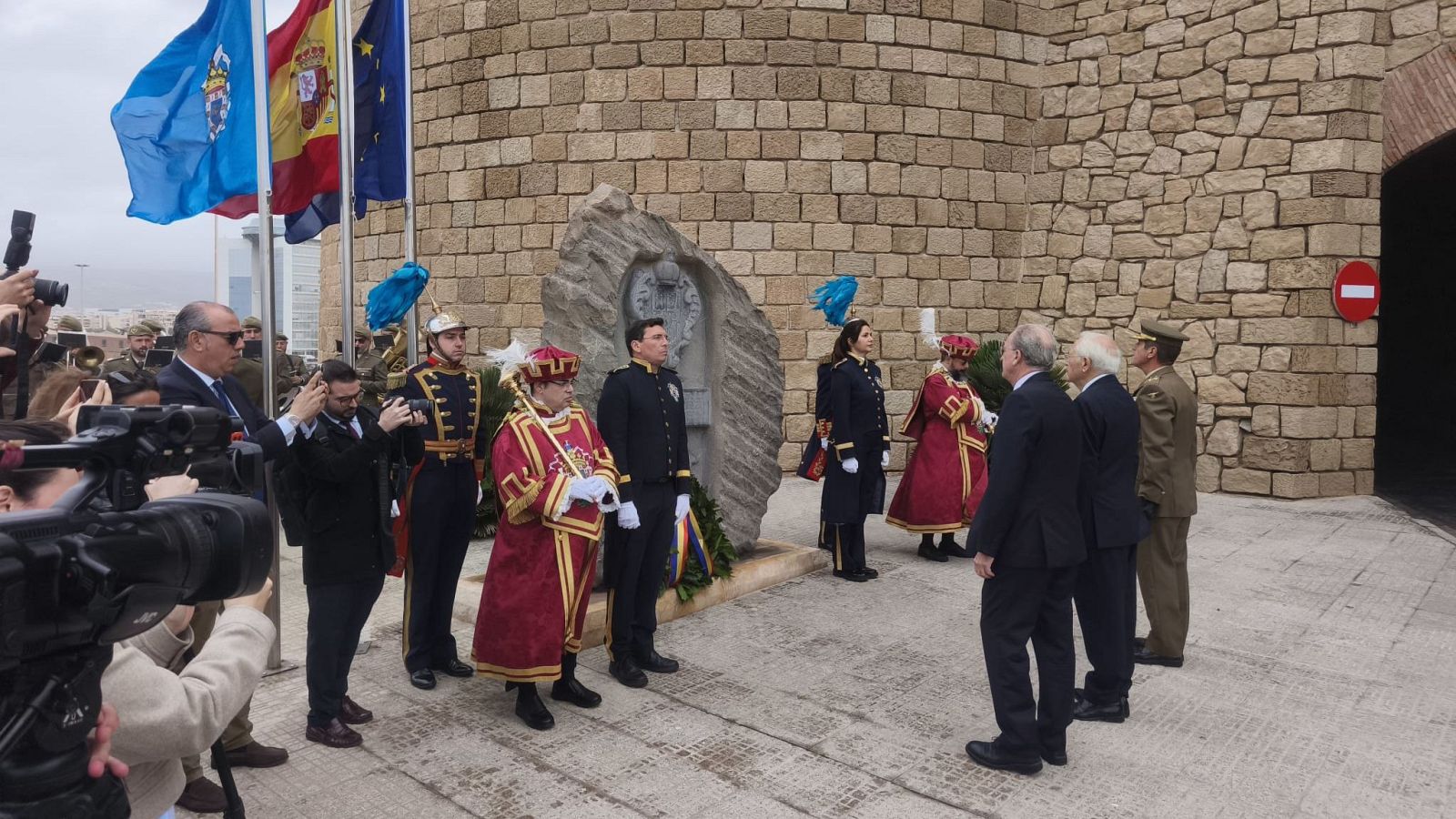 Una ceremonia conmemorativa muestra una ofrenda floral frente a una estructura de piedra, con personas vestidas formalmente y con uniformes militares, algunas con sombreros con plumas, mientras se observan banderas y equipos de grabación.