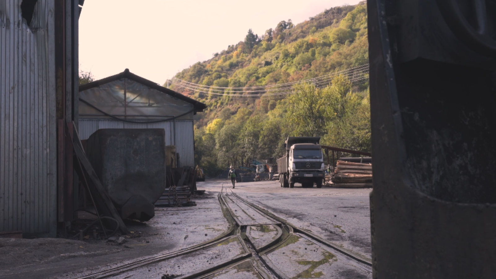 En la imagen se observa un entorno industrial con vías de tren que se bifurcan, un camión estacionado y una persona caminando. Al fondo, se aprecia una colina cubierta de vegetación, sugiriendo un lugar de trabajo.
