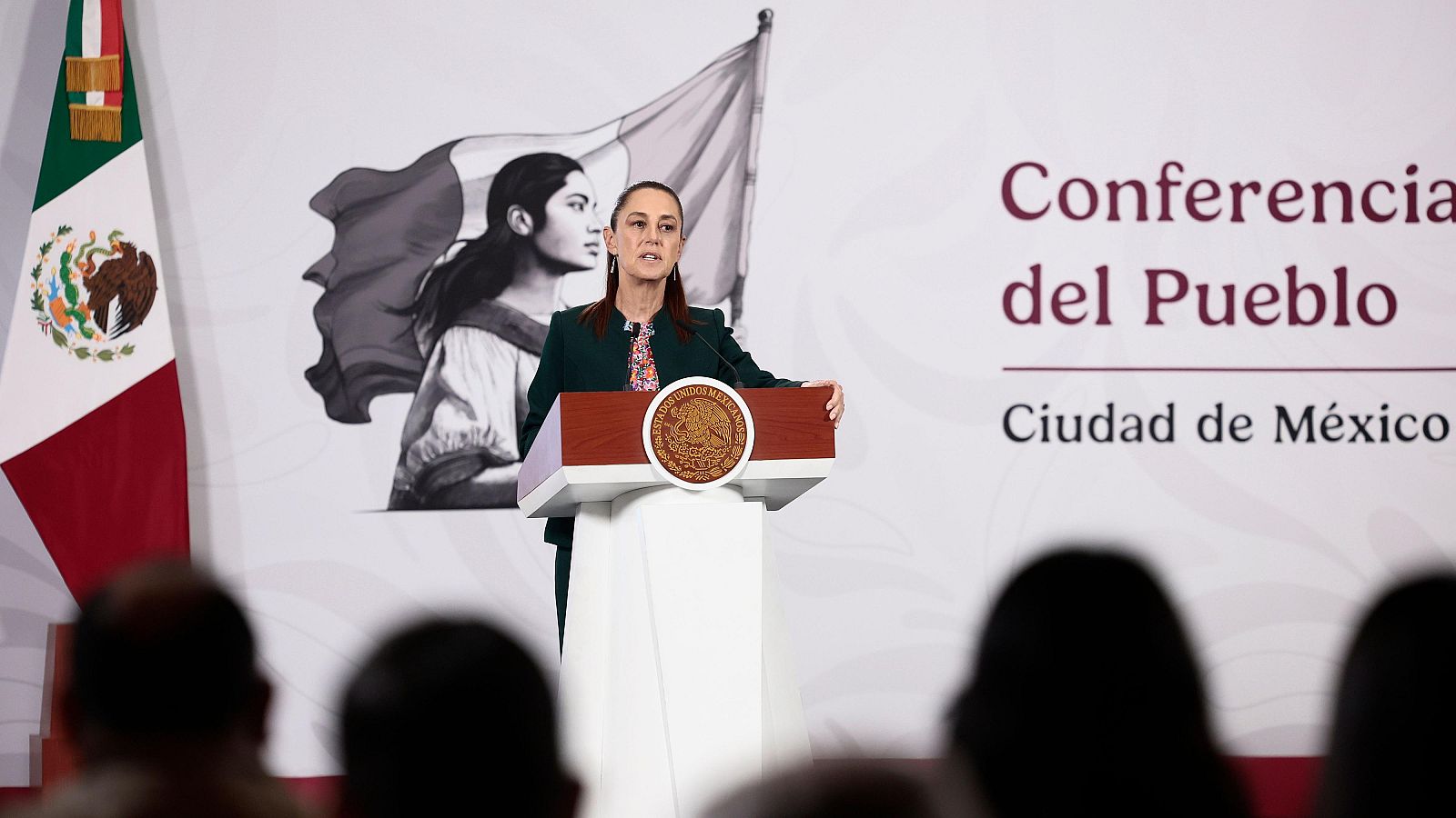 En el Palacio Nacional de la Ciudad de México, la presidenta de México, Claudia Sheinbaum, ofrece una conferencia de prensa. Se encuentra detrás de un atril con el escudo nacional, vestida con una chaqueta oscura, con una bandera mexicana y una imagen en blanco y negro de fondo.