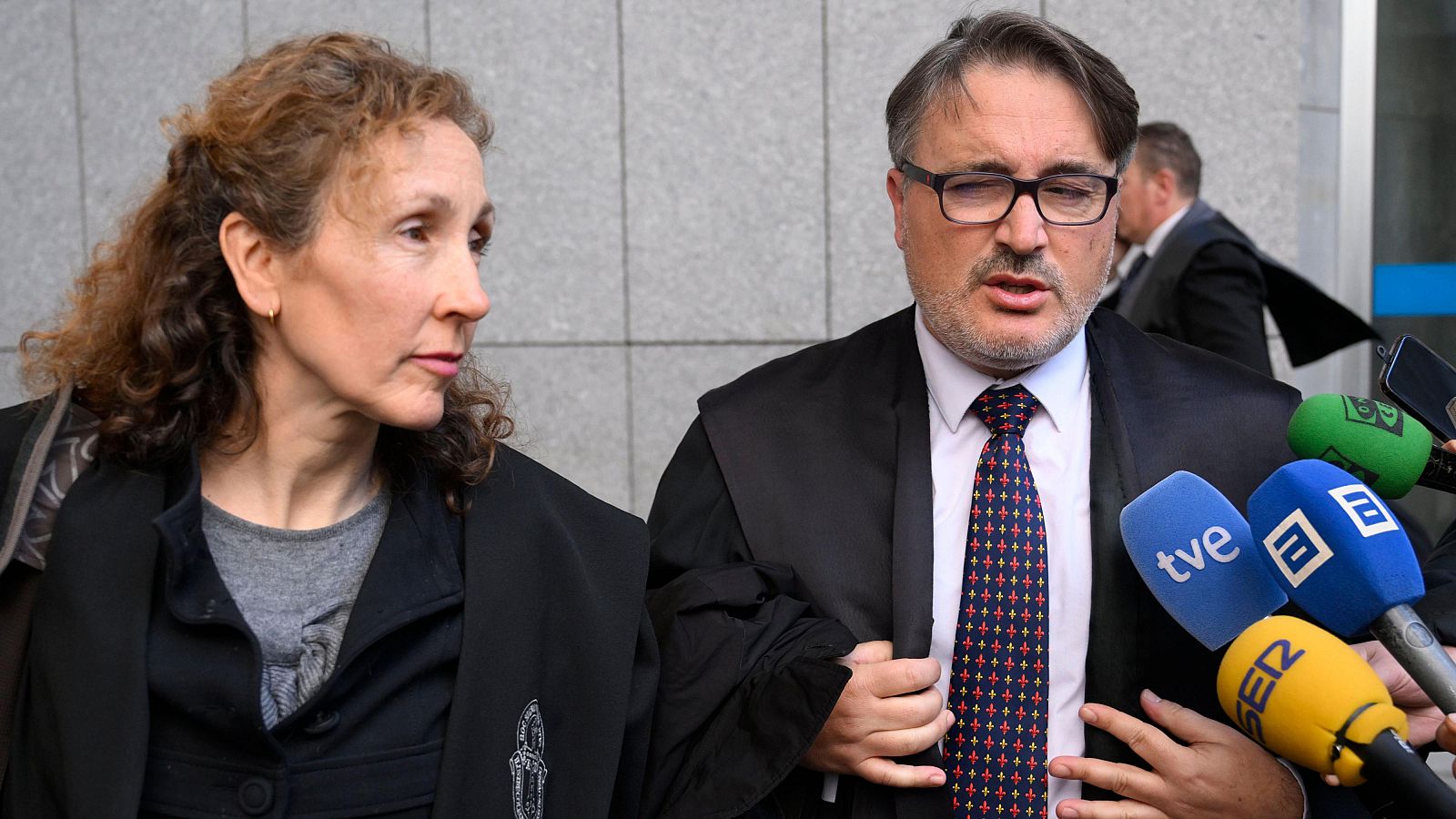 Dos abogados, Javier Muñoz y Elena González, posan para la cámara. Elena, con el pelo rizado, lleva una blusa gris y chaqueta negra. Javier, con gafas y traje, está rodeado de micrófonos.