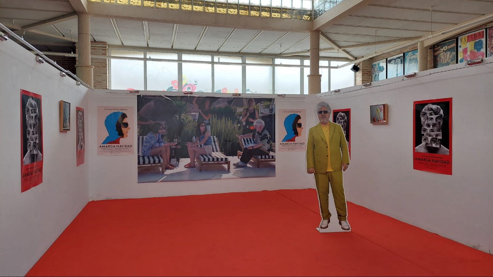 En un photocall de un festival de cine, una figura de cartón y carteles promocionan una película. El espacio, con alfombra roja, está iluminado por luz natural y presenta una lona con una escena del filme.