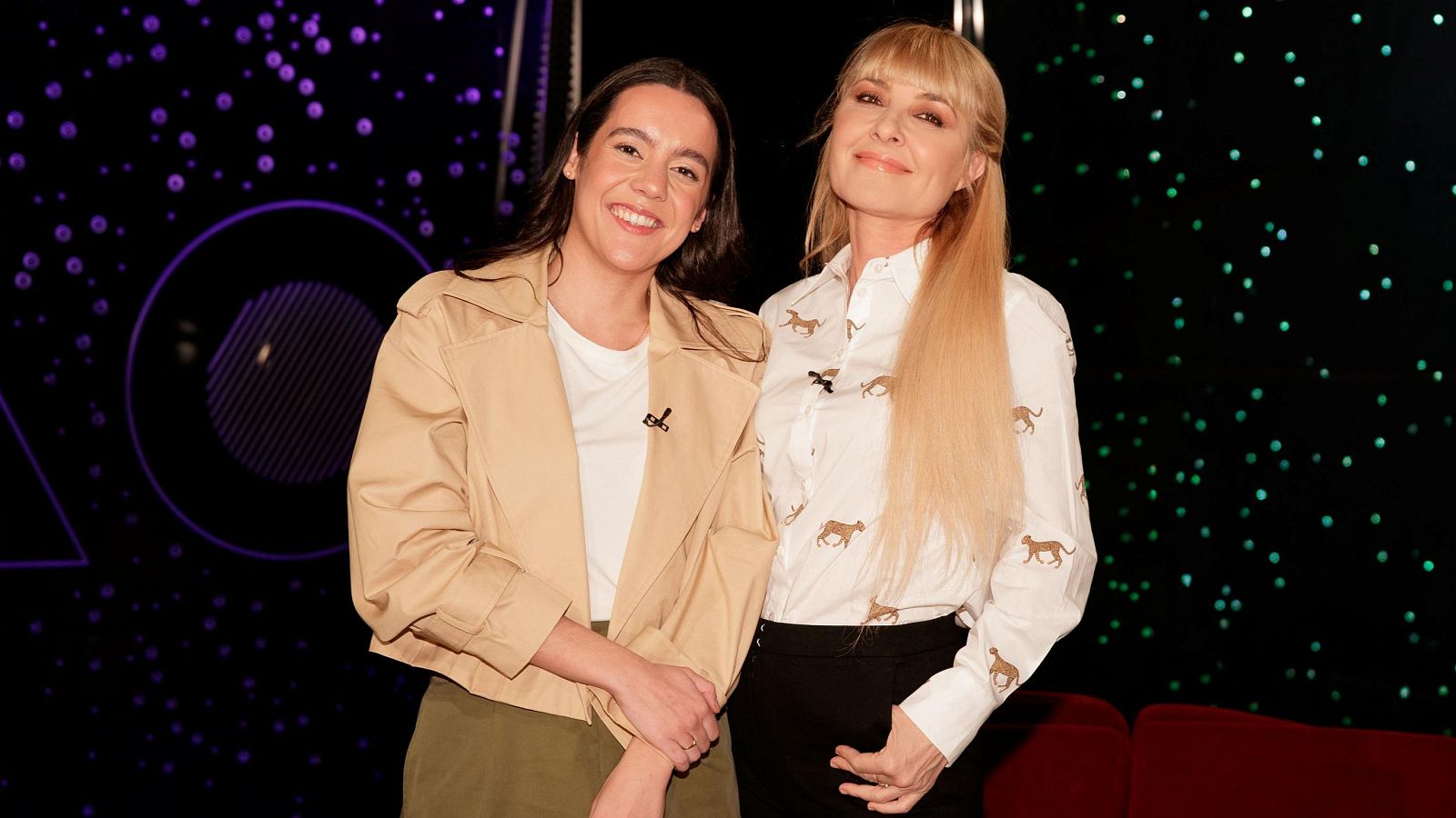 Dos mujeres sonríen juntas en una imagen. Una de ellas, con pelo oscuro, lleva una camiseta blanca y chaqueta beige; la otra, con pelo rubio, viste una camisa estampada. El fondo es oscuro con luces de colores.