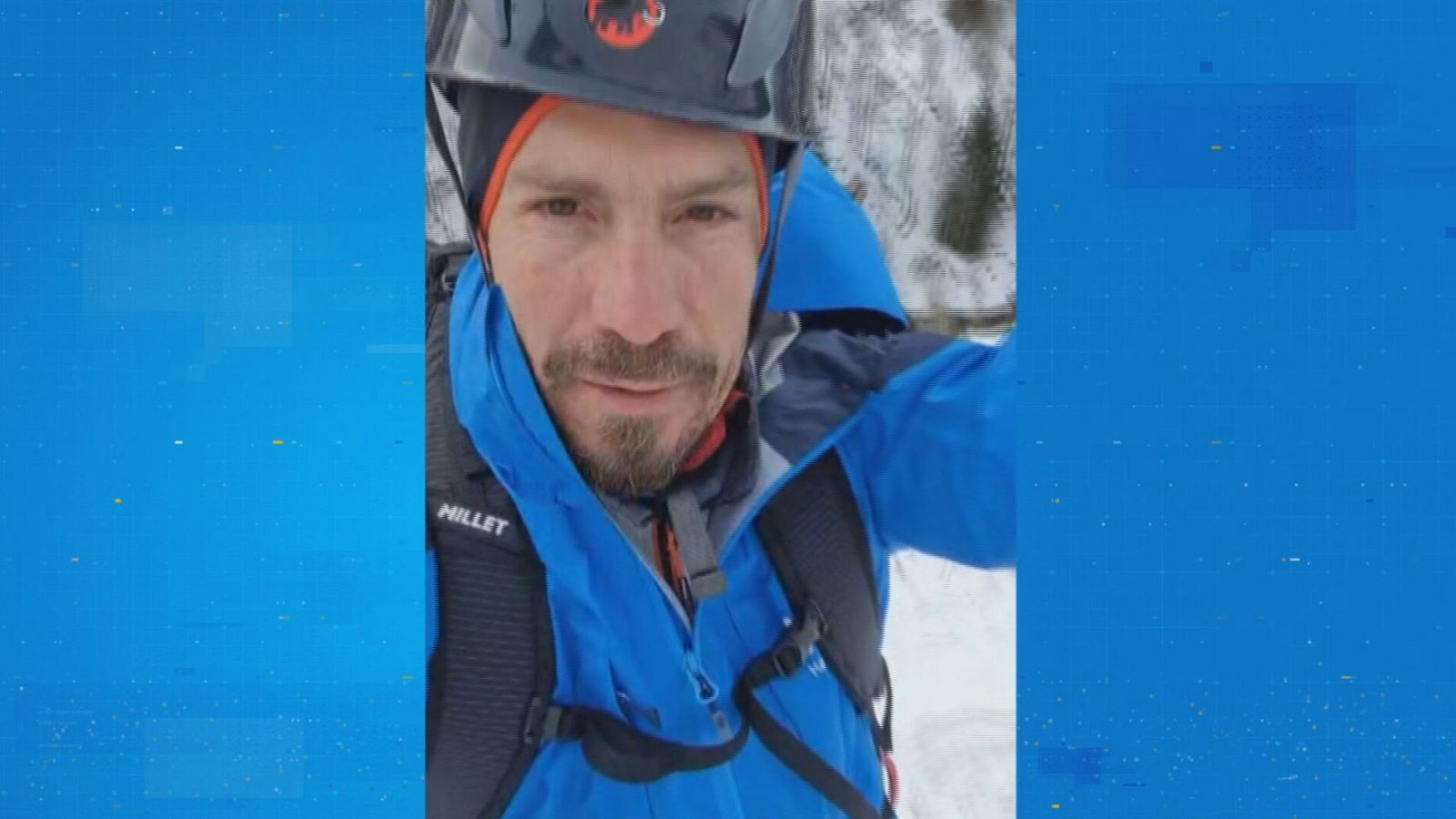 Un hombre con barba y bigote, equipado con casco y ropa de montaña, posa frente a un paisaje nevado. El deportista lleva un casco negro con el logo de una marca y una chaqueta azul.