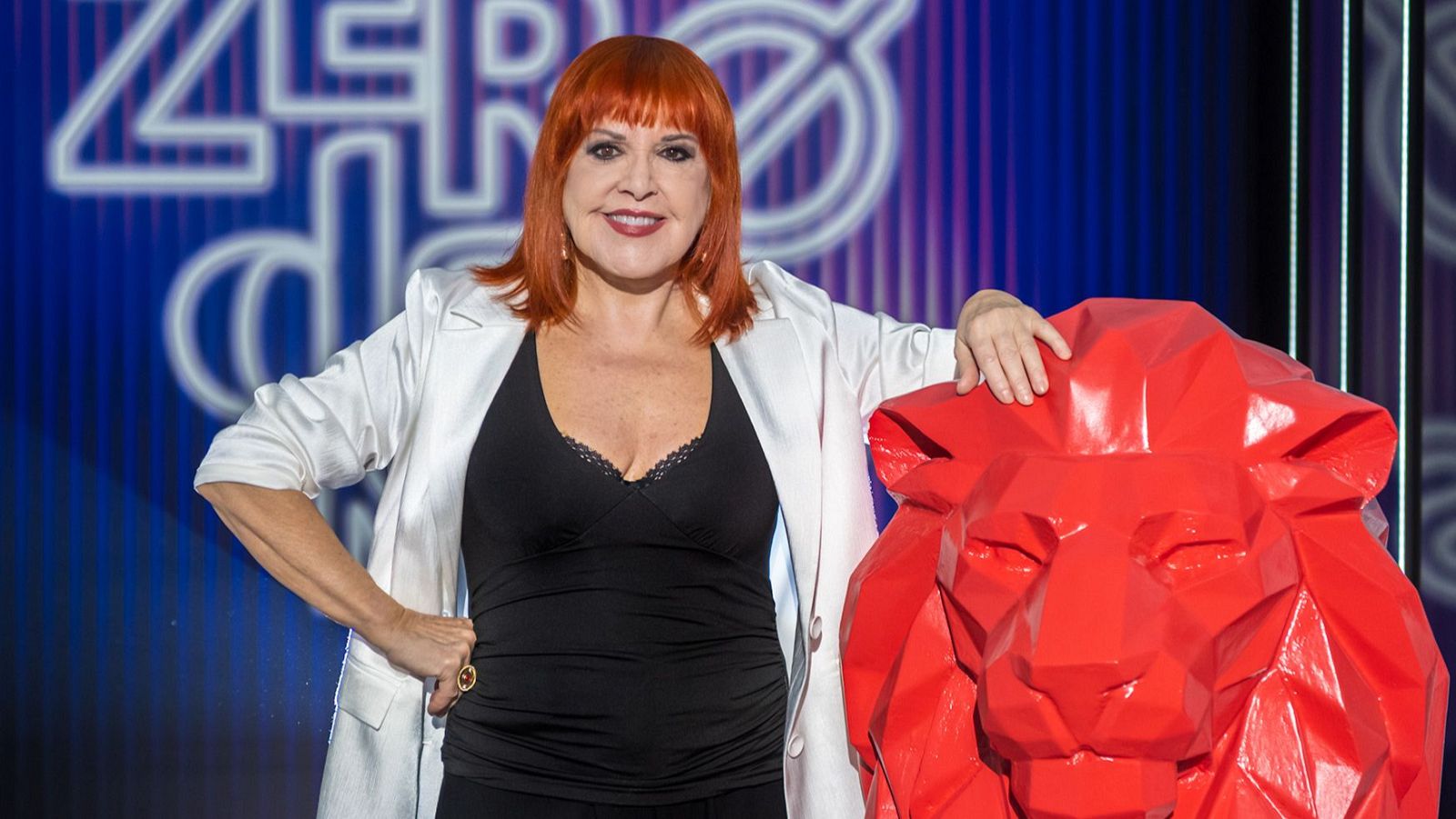 Una mujer pelirroja sonríe, vestida con una camiseta negra y chaqueta blanca, junto a una escultura roja con forma de león, sobre un fondo azul con líneas verticales blancas.