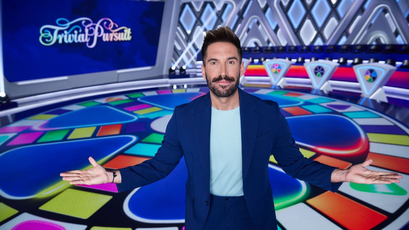 En el plató de un programa de televisión, un hombre con barba y chaqueta azul posa frente a un tablero circular con casillas de colores. El presentador, con el pelo peinado hacia atrás, sonríe a la cámara.