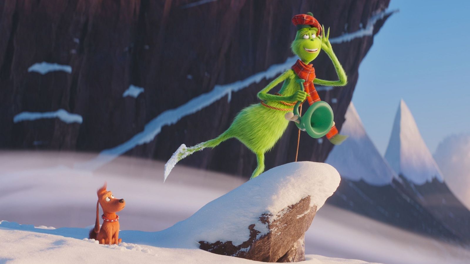 El Grinch, con gorro y bufanda roja, se alza sobre una roca nevada, sosteniendo un objeto verde. A su lado, en la nieve, se encuentra un perro con collar rojo, en un paisaje montañoso y nevado.