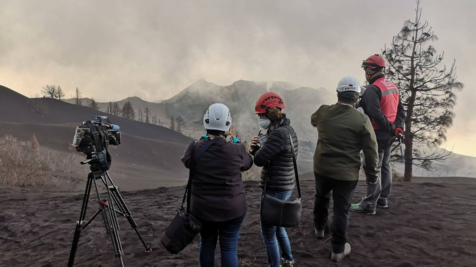 En un entorno volcánico desolado, un equipo de filmación, con cascos y un chaleco con siglas, prepara su equipo frente a un volcán humeante, posiblemente para un reportaje.
