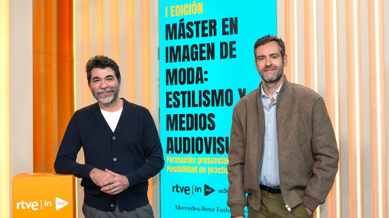 Dos hombres posan frente a un cartel promocional. Uno de ellos, con barba canosa y cárdigan azul oscuro, está junto a otro con barba incipiente y chaqueta marrón. Detrás, un cartel azul anuncia un máster en imagen de moda, con logos de RTVE y la universidad.