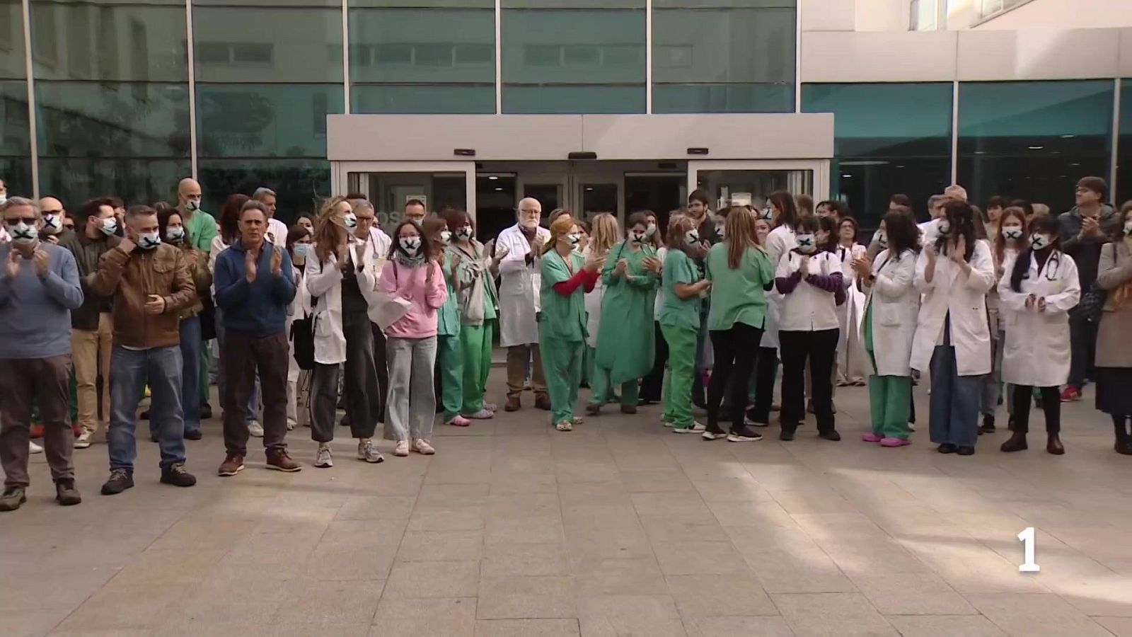 Un grupo de personas, probablemente personal sanitario, se manifiesta frente a un hospital. Todos llevan mascarillas con una cruz negra y aplauden, posiblemente en señal de protesta.