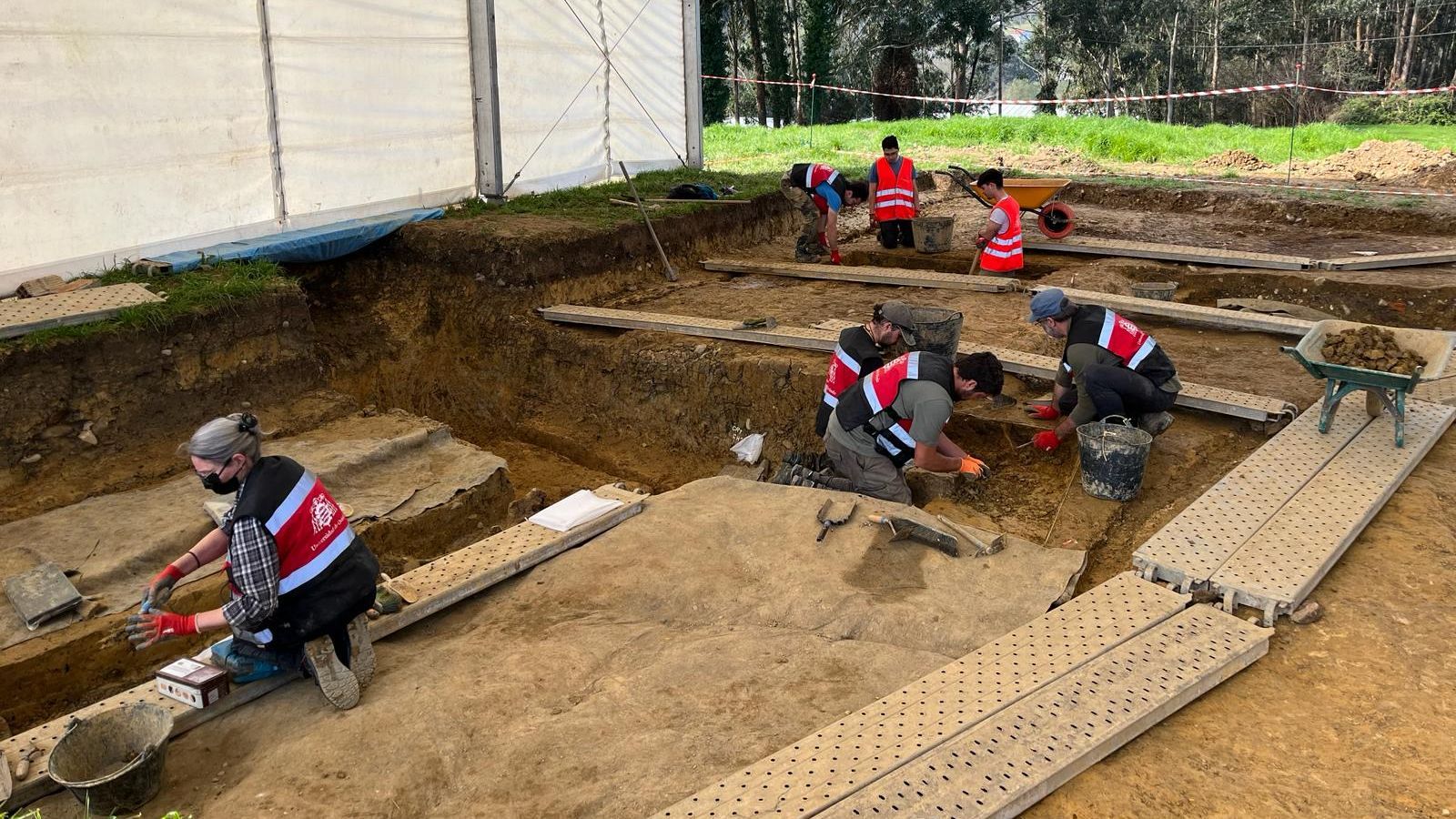 En una excavación al aire libre, un grupo de personas trabaja en fosas, utilizando herramientas manuales y llevando chalecos reflectantes. Se observa una estructura de lona y vegetación en el entorno.
