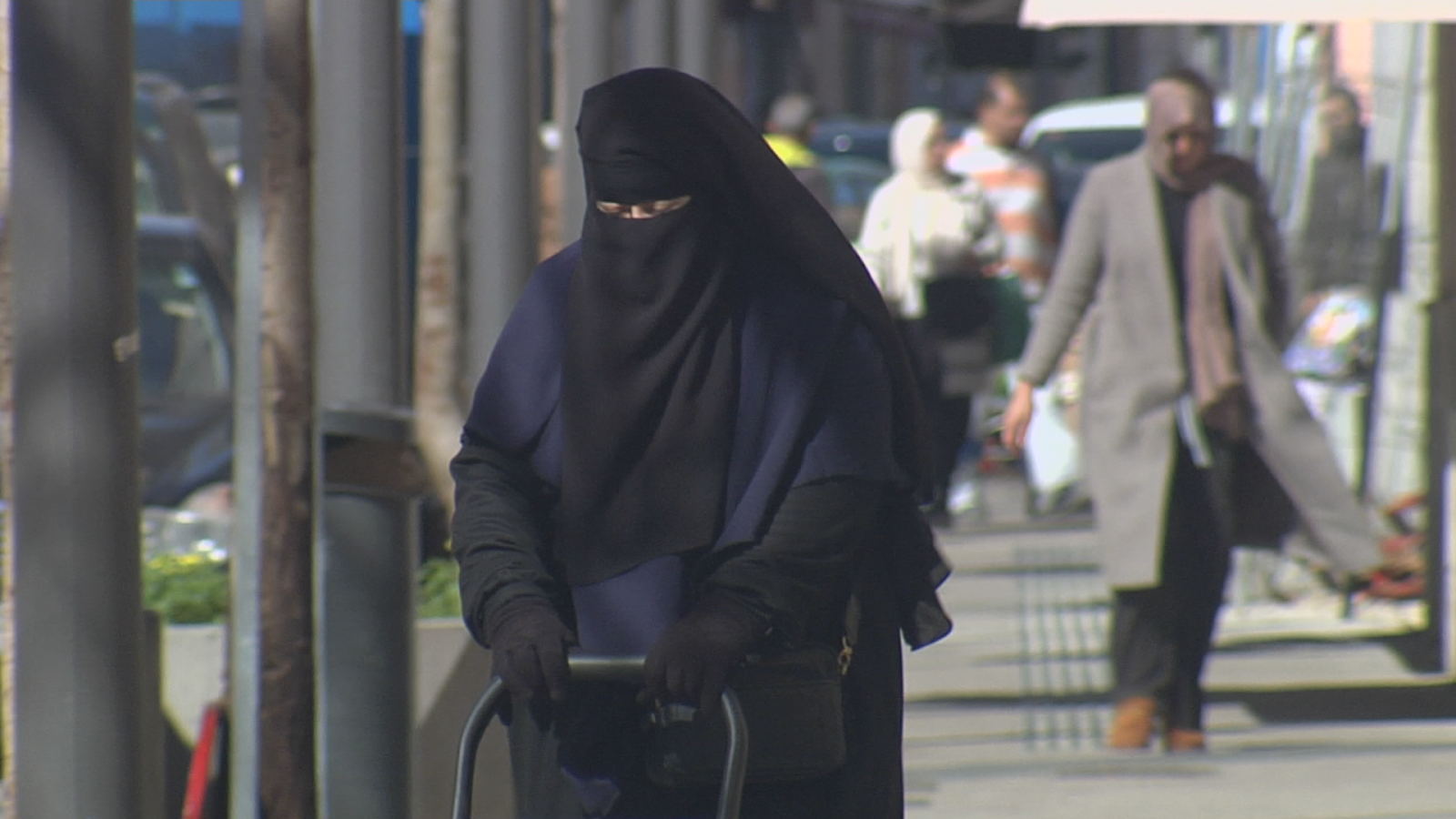 Una mujer con burka negro camina por una calle urbana, empujando un carrito de la compra. La vestimenta cubre todo su cuerpo, con excepción de los ojos.