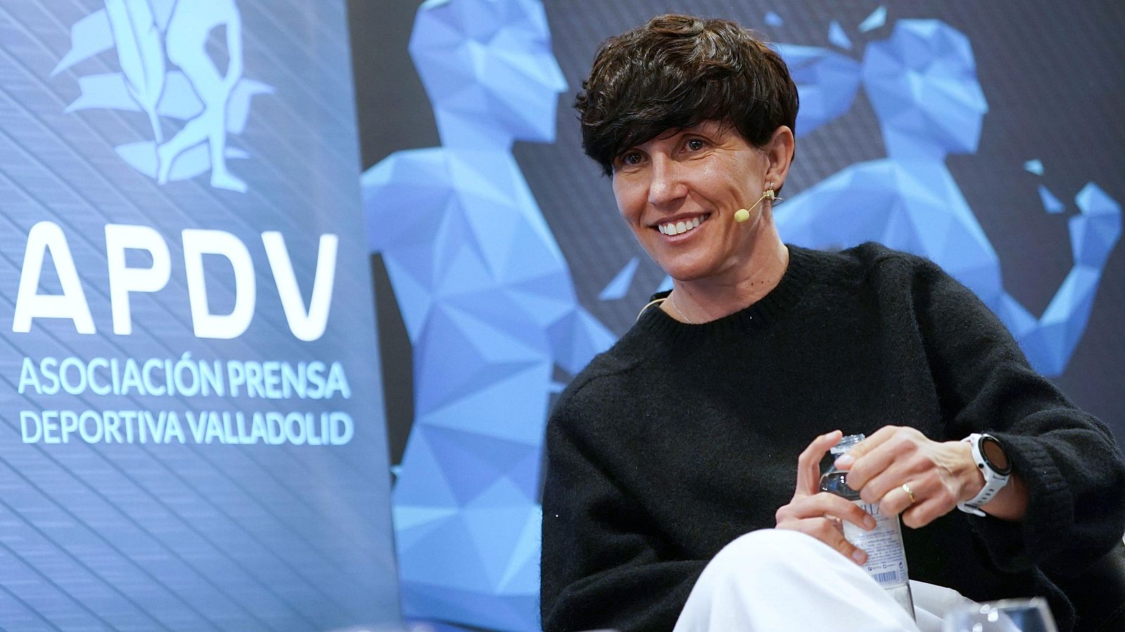 Una mujer sonriente, con un micrófono y un reloj, posa con una botella de agua. Detrás de ella, se distingue un logotipo.