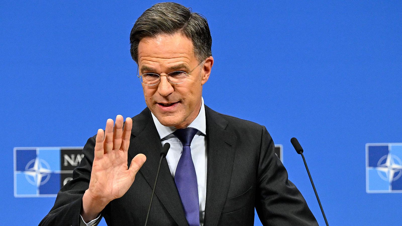 Mark Rutte, con traje oscuro y corbata azul, habla frente a un micrófono, gesticulando con la mano levantada. Detrás de él, se observa un panel azul con un logo.