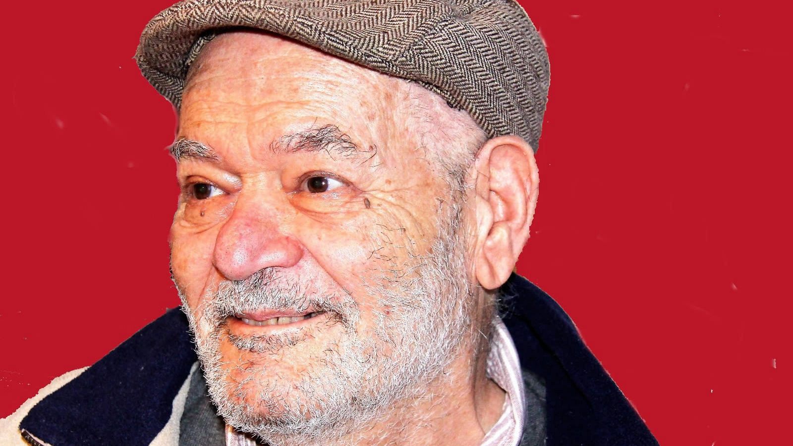 Retrato de un hombre mayor con cabello y barba canosos, usando una gorra de tela con diseño de espiga y una chaqueta oscura sobre una camisa blanca, con un fondo rojo.