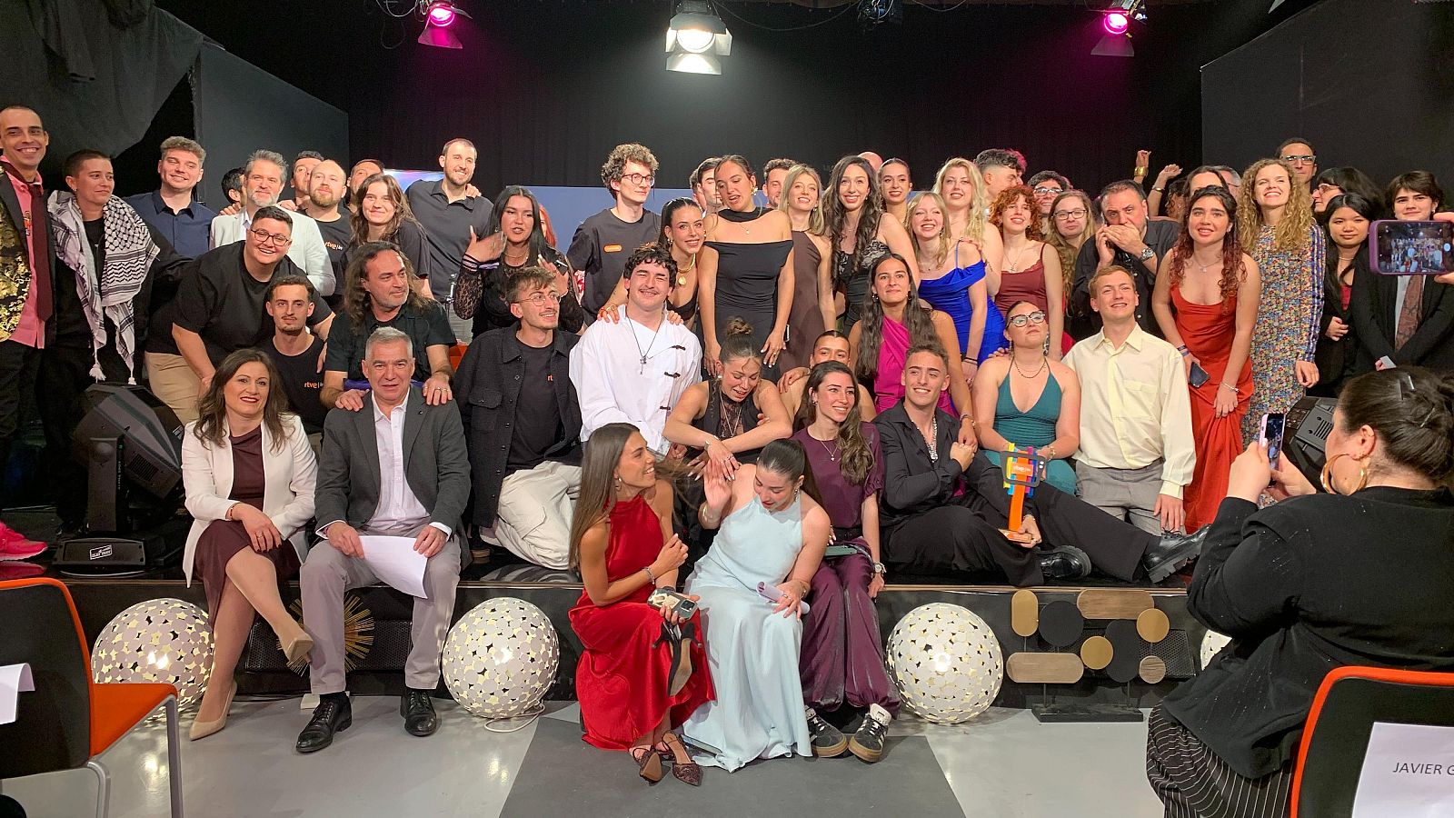 En un escenario iluminado y decorado con esferas, un grupo numeroso posa para una foto durante un evento. Se distinguen personas vestidas de manera formal y casual, con alguien fotografiando la escena con un móvil.