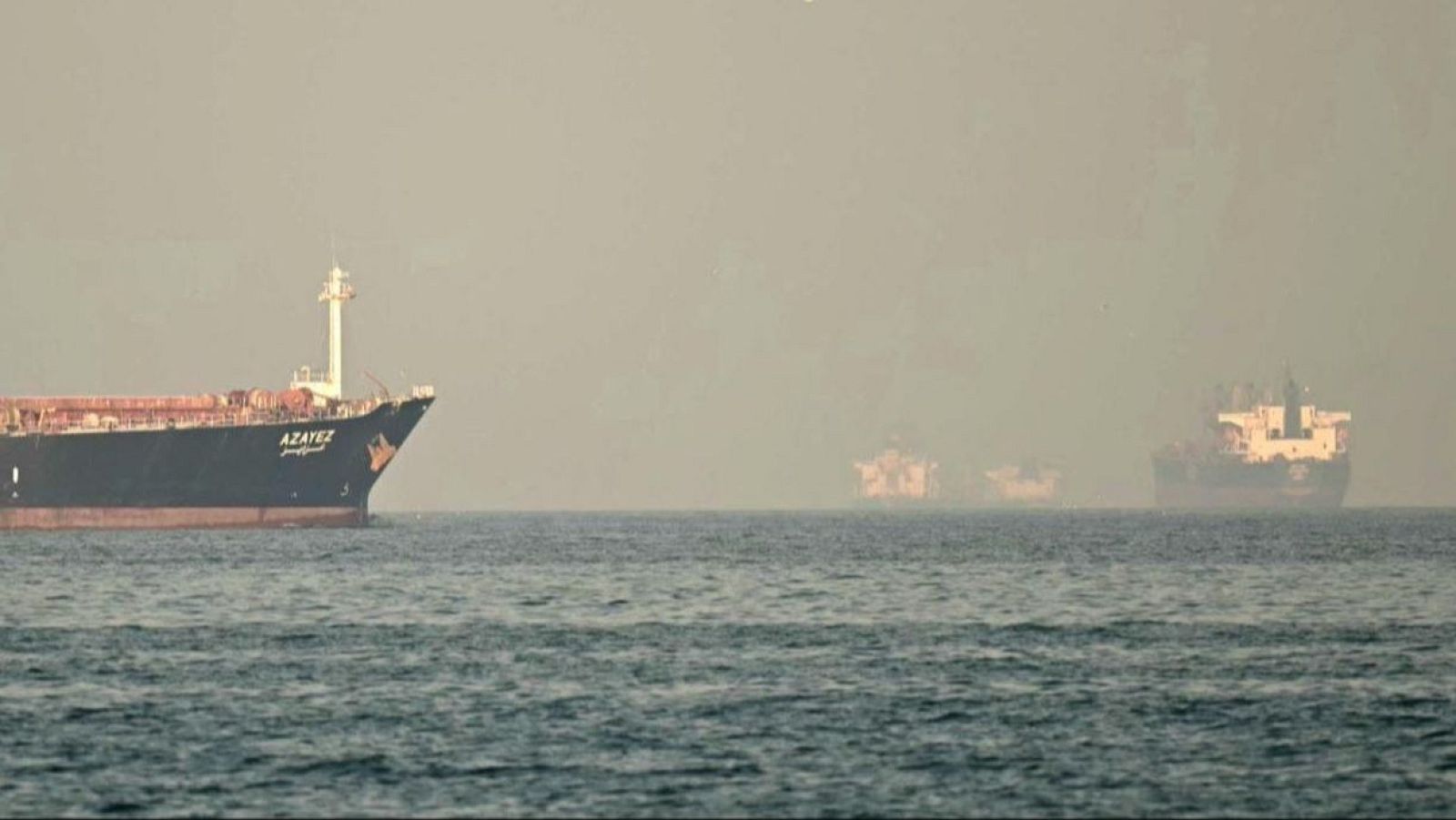 En un mar brumoso, un barco oscuro con la palabra "AZAYEZ" en su casco navega, acompañado por otros barcos menos visibles debido a la niebla, con el horizonte difuminado por la misma.