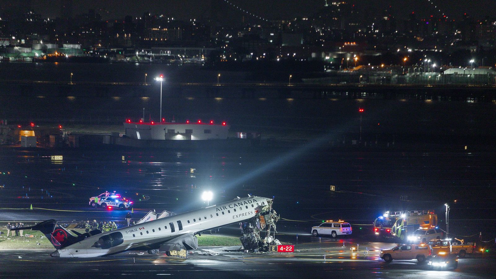 Un avión dañado de Air Canada Express está en una pista de aeropuerto por la noche, rodeado de vehículos de emergencia con luces. La parte frontal del avión presenta daños considerables, y al fondo se observa una ciudad iluminada.