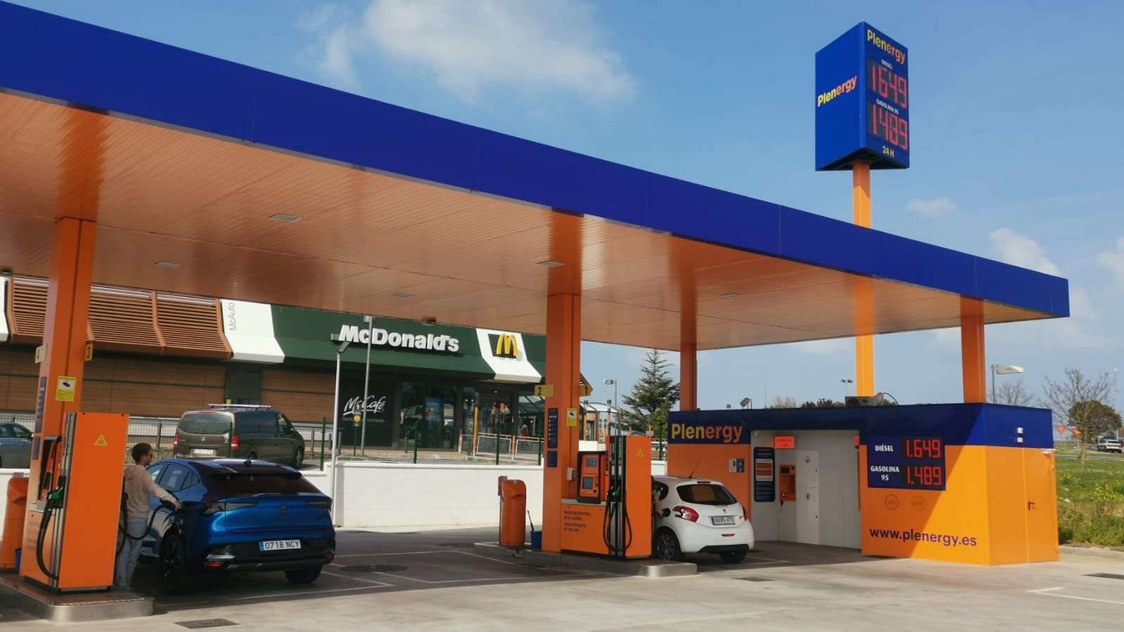 Dos coches repostan combustible en una gasolinera, uno azul y otro blanco. Un empleado está manipulando la manguera. Se pueden ver los precios del combustible en un tótem.