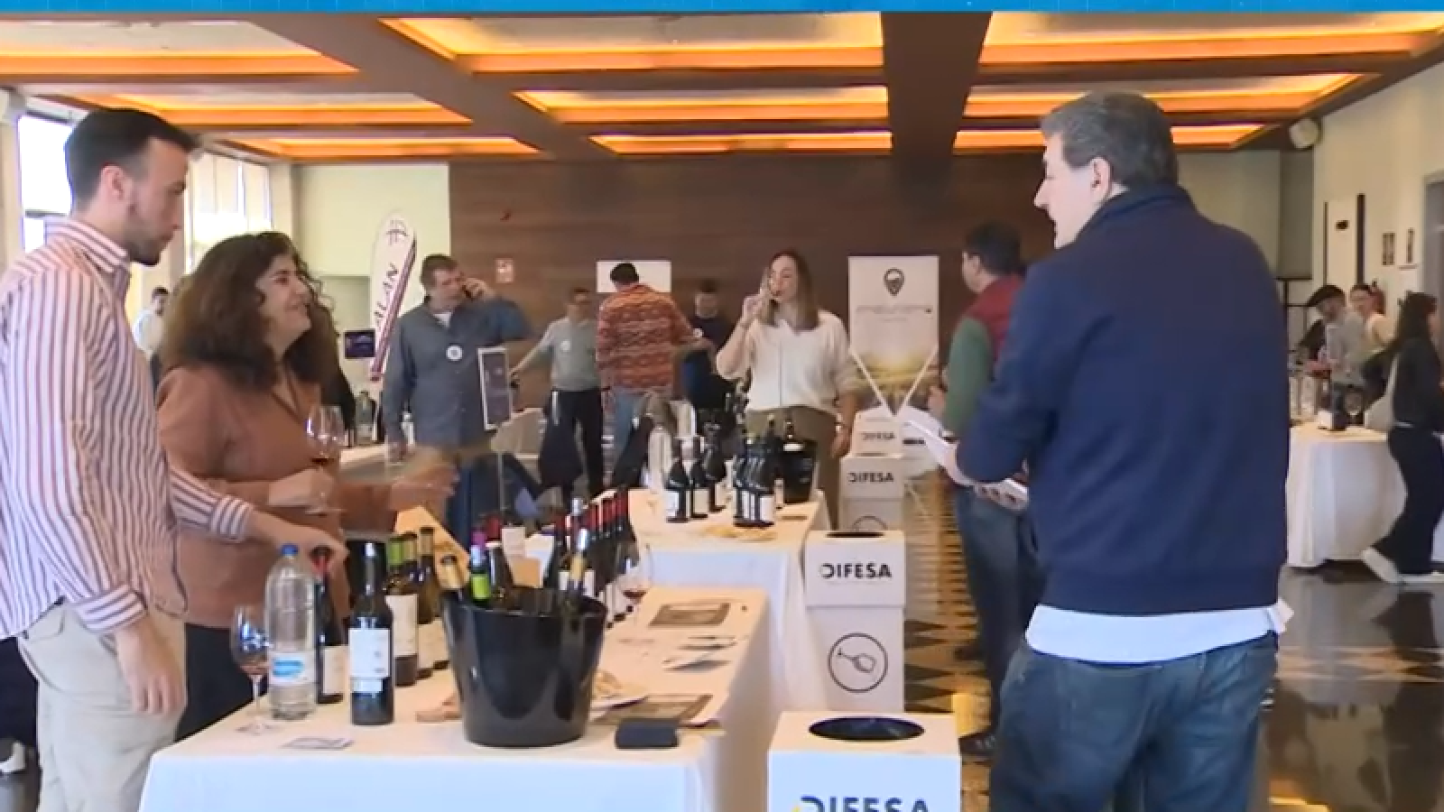 En la imagen se observa una reunión social con mesas vestidas con manteles blancos, botellas de vino, copas y cubiteras. Varias personas están reunidas, posiblemente degustando vino y conversando.