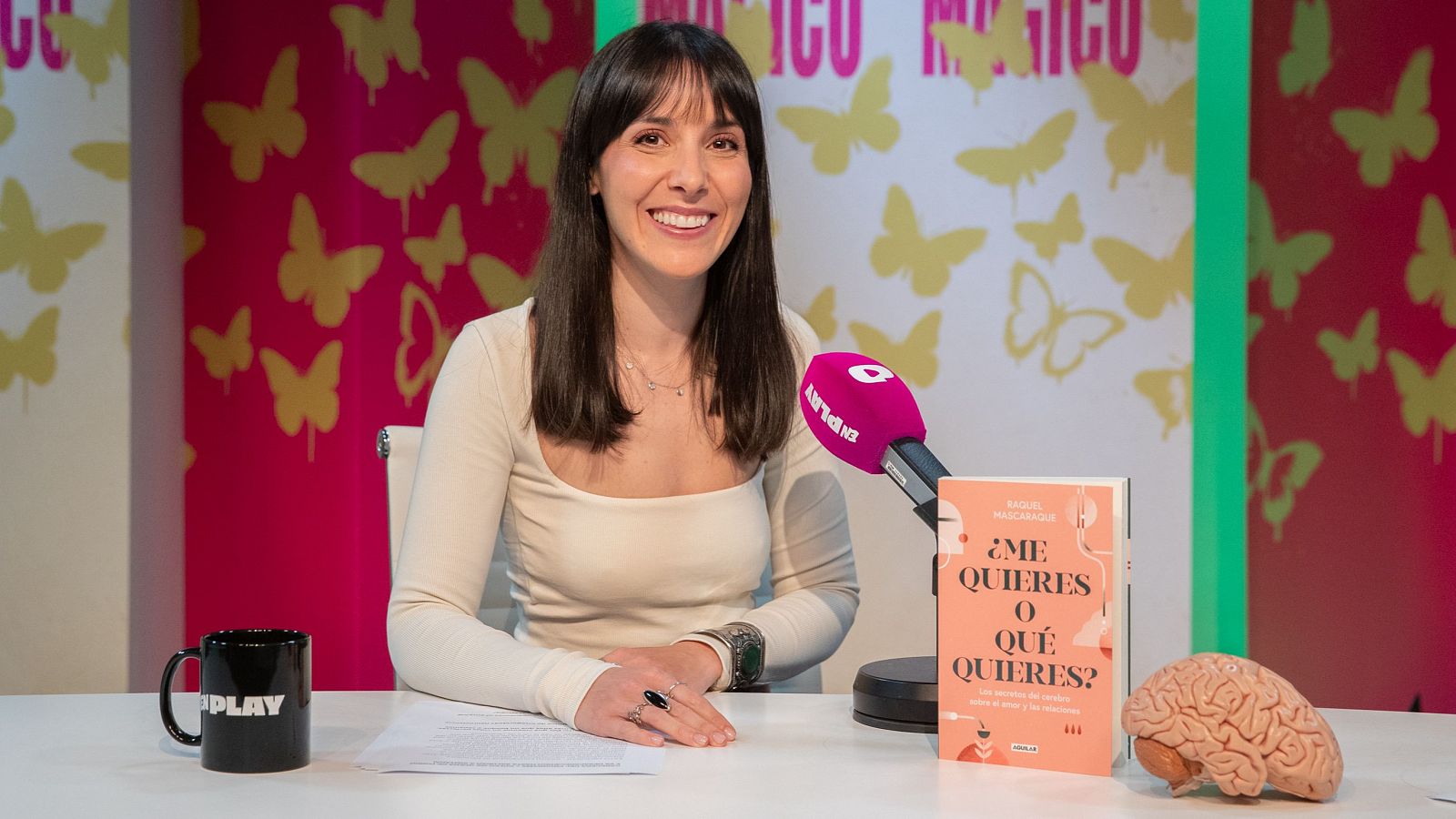 En la imagen, una mujer sonriente con blusa crema y cabello castaño oscuro posa junto a un libro y un modelo de cerebro. Una taza negra con la palabra "PLAY" y un micrófono rosa complementan la escena, con un fondo decorado con mariposas.