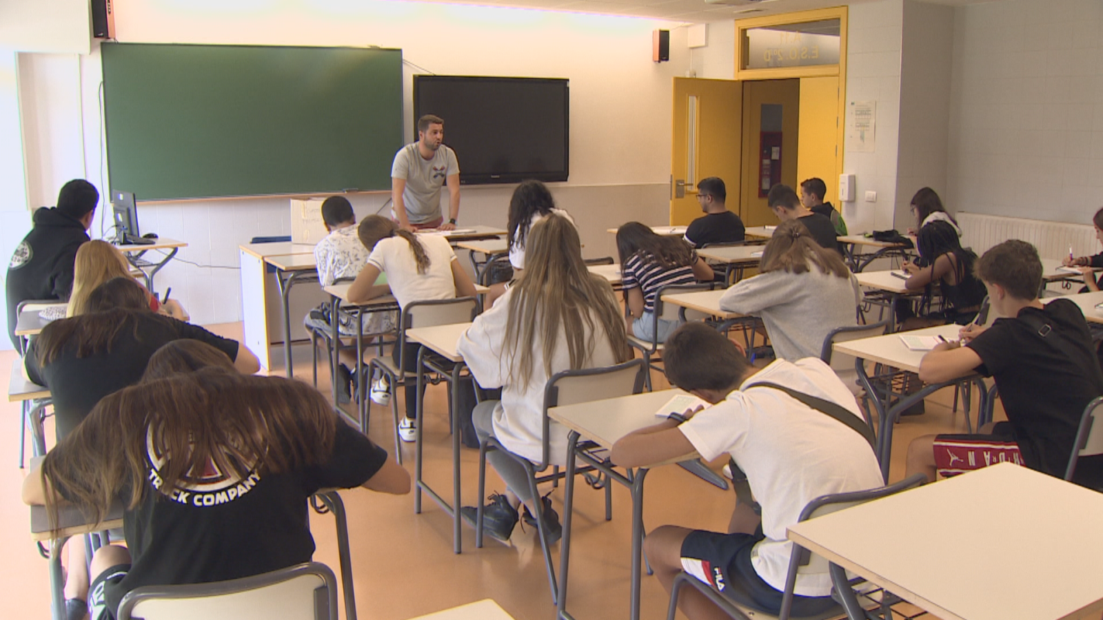En un aula, estudiantes sentados en pupitres individuales escriben mientras un profesor está de pie frente a una pizarra. Se observa una puerta amarilla al fondo.