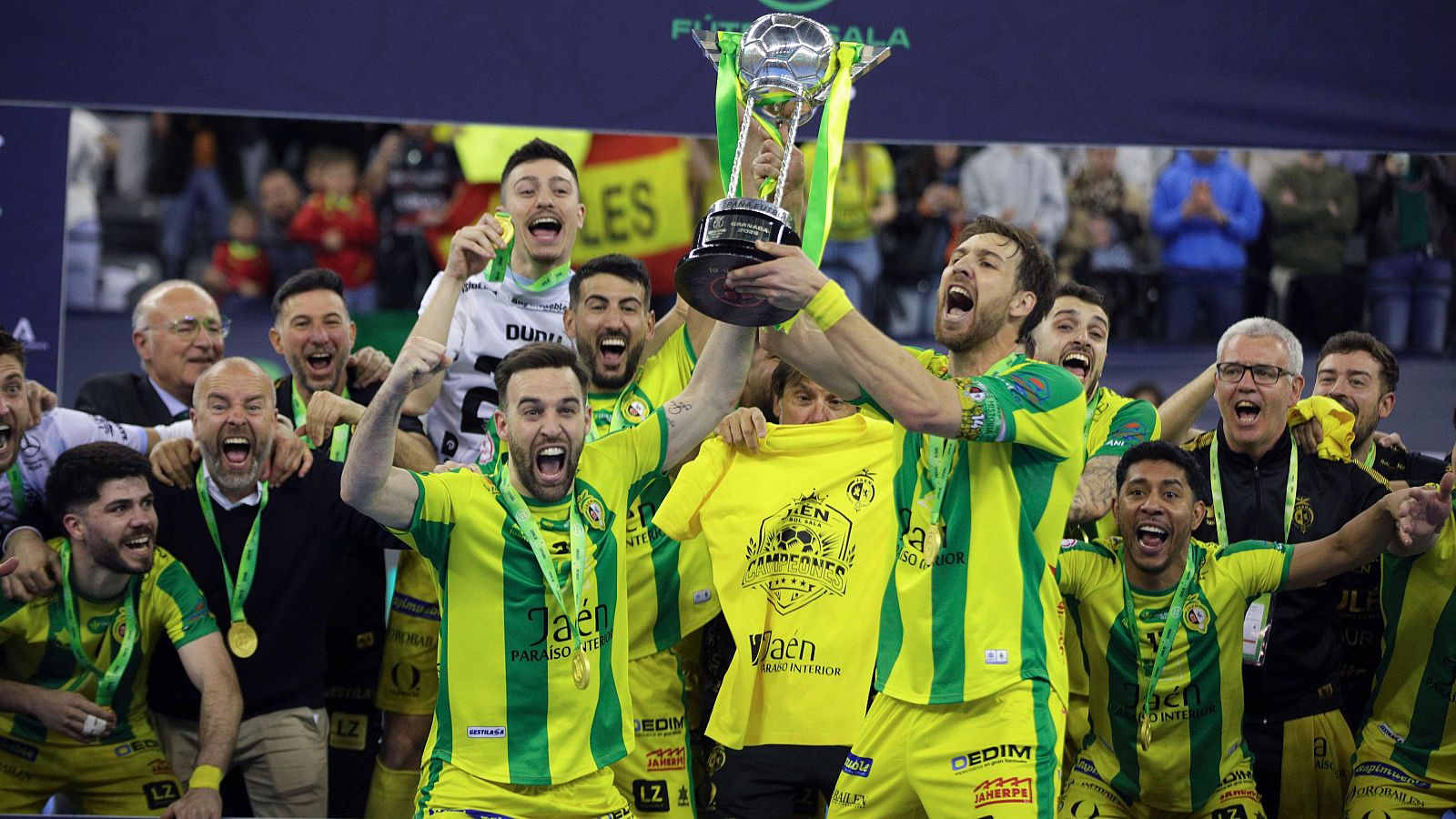 Un grupo de jugadores de fútbol sala, vestidos con camisetas amarillas y verdes, celebran la victoria levantando una copa plateada, con un ambiente de euforia y alegría.