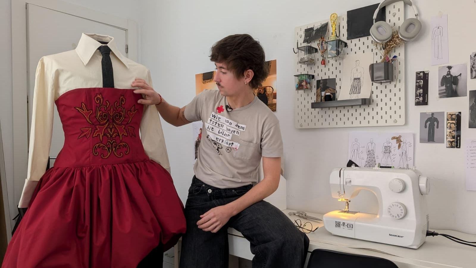 Un joven con bigote y aretes, vestido con camiseta gris y pantalones vaqueros, posa junto a un maniquí que luce una camisa blanca con corbata y un conjunto rojo con detalles dorados. En el fondo, una pared blanca con elementos de diseño.
