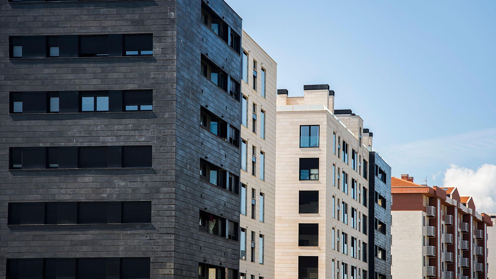 Una fila de edificios modernos de diferentes alturas y estilos arquitectónicos se alza bajo un cielo despejado. Se observa un contraste entre edificios oscuros y claros, con ventanas de diversos tamaños y diseños.
