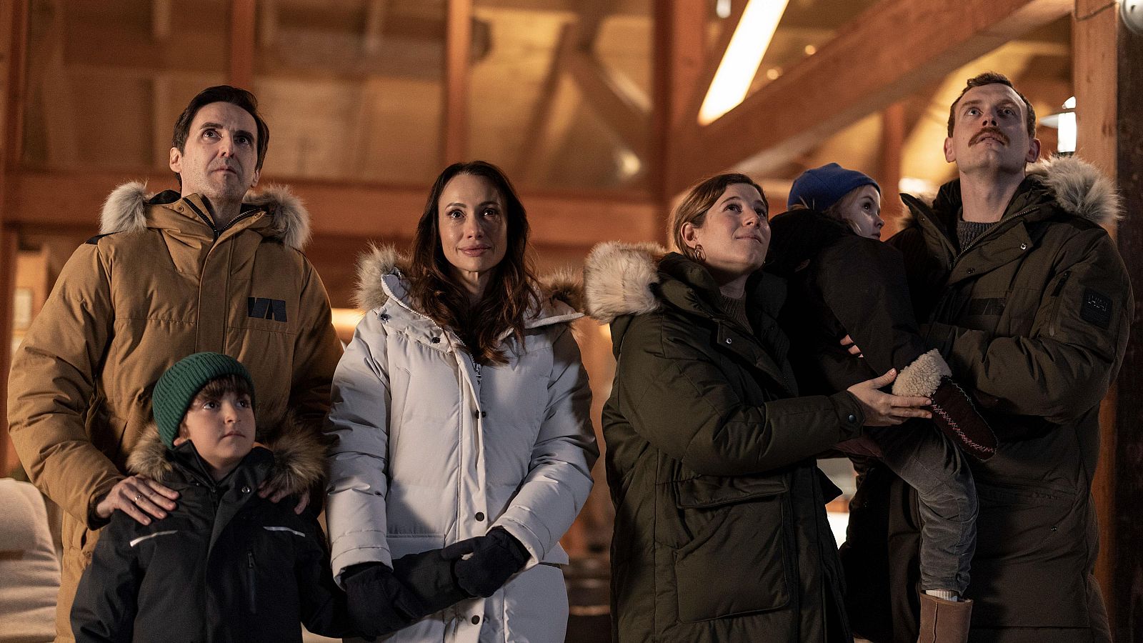 Un grupo familiar, compuesto por dos parejas y dos niños, observa algo fuera de la imagen. Los adultos visten chaquetas de invierno y los niños gorros, en un entorno con techo de madera, sugiriendo un ambiente frío y familiar.