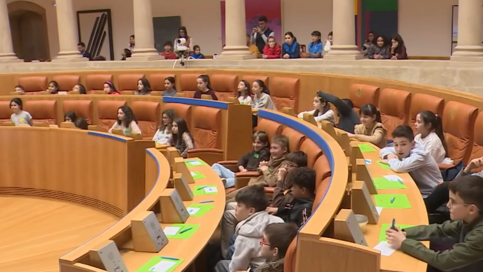 En el parlamento, un grupo numeroso de niños se encuentra sentado en los asientos, que tienen mesas con papeles. Algunos niños están escribiendo o dibujando, mientras que adultos observan desde la galería superior.