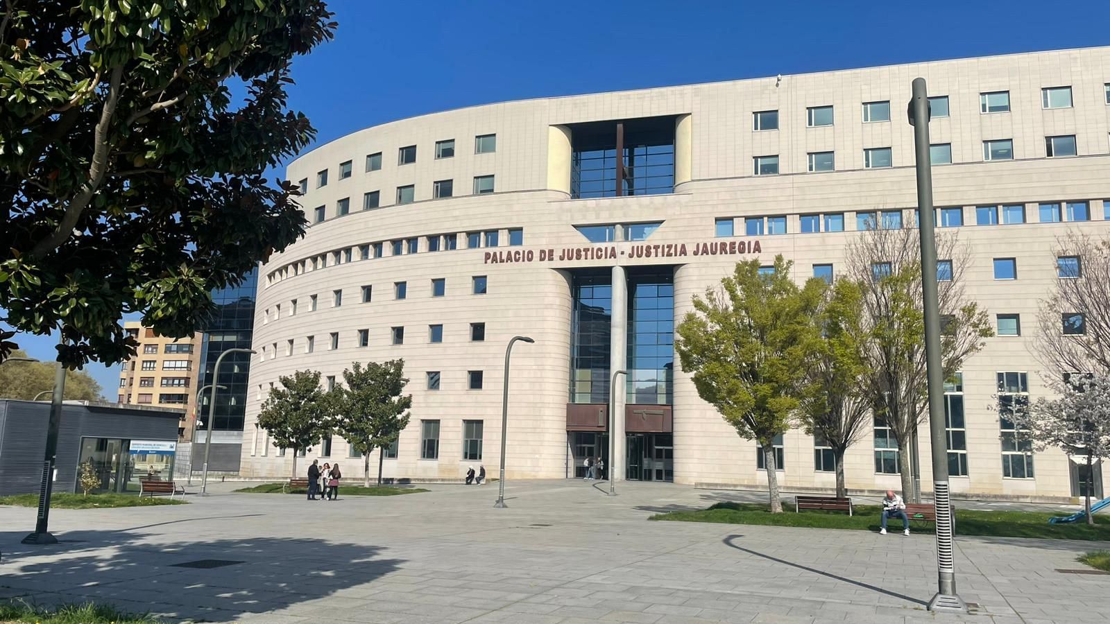 Fachada del Palacio de Justicia de Navarra, de estilo moderno, con una inscripción en la parte superior y una plaza con árboles y personas en la parte delantera.