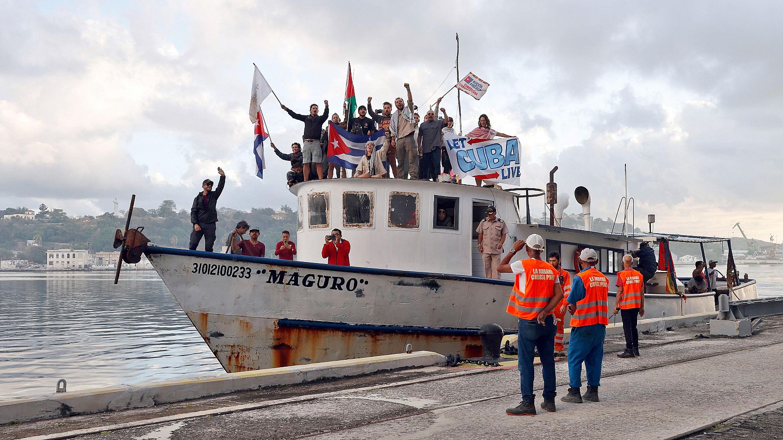 Un barco blanco, el "MAGURO", llega a un puerto cubano, donde un grupo de personas celebra con banderas y una pancarta. En el muelle, individuos con chalecos naranjas observan la escena de la llegada.