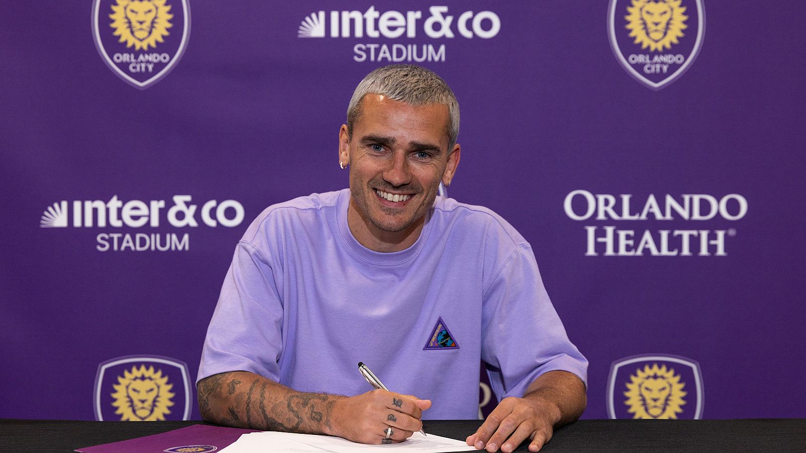 El futbolista Antoine Griezmann, con el pelo corto y una camiseta lila, firma un contrato. Se aprecian tatuajes en sus brazos y sonríe a la cámara, con logos en el fondo.